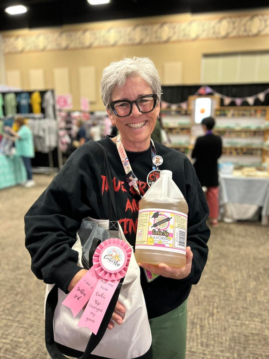 A smiling person with glasses holding a plastic jug and a pink ribbon at an indoor craft or trade show event.