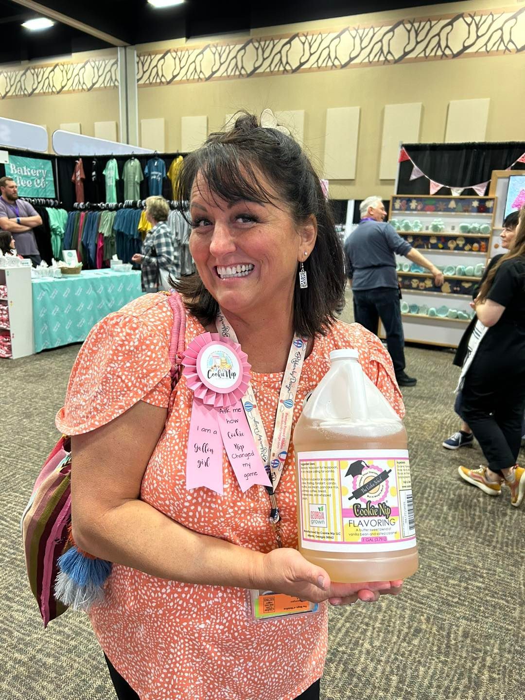 A person smiling while holding a large plastic jug of liquid at an indoor craft show or convention.