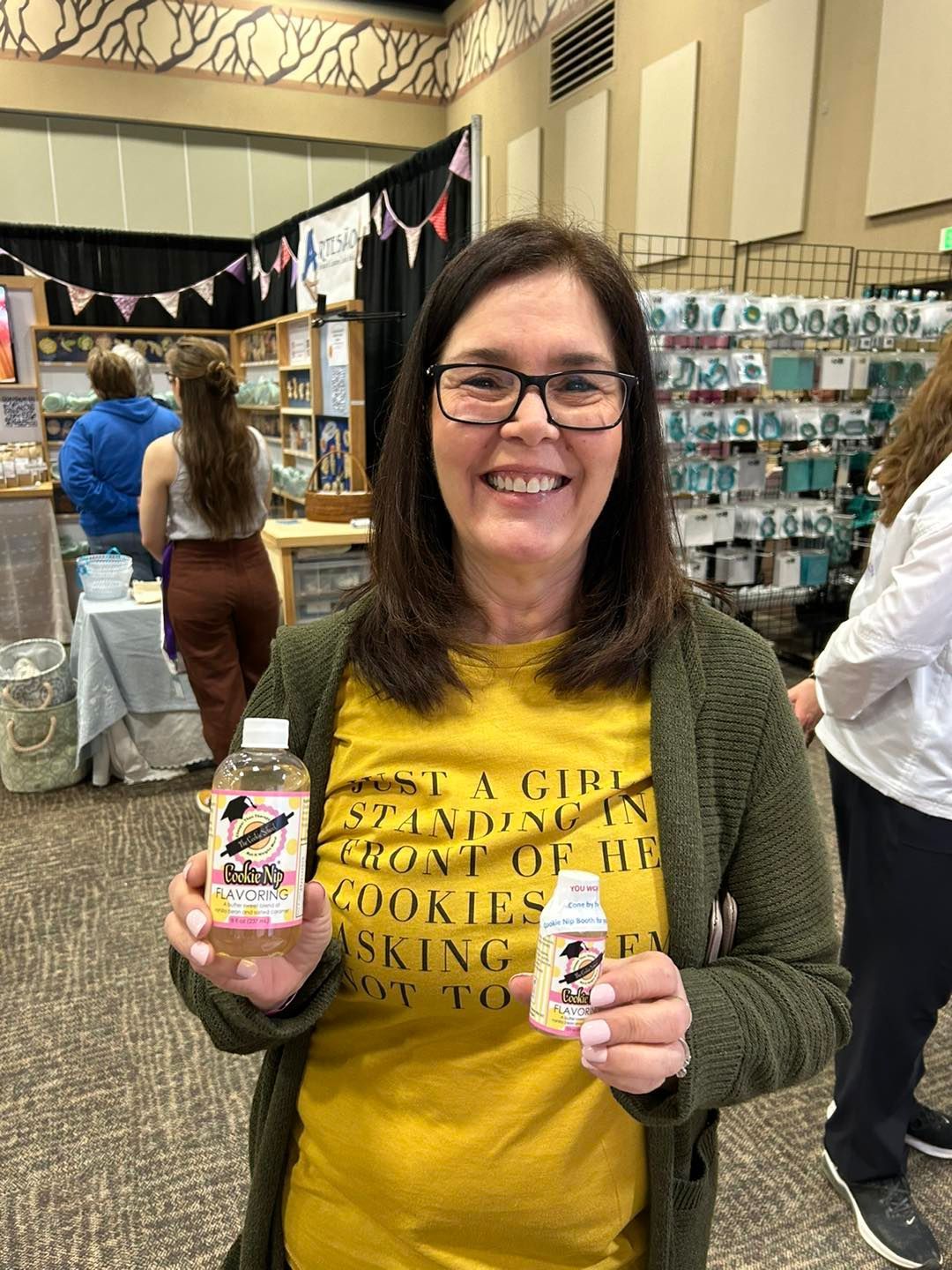 A smiling person in a yellow graphic t-shirt and green cardigan holds two bottles of Cookie Nip at an indoor craft fair.
