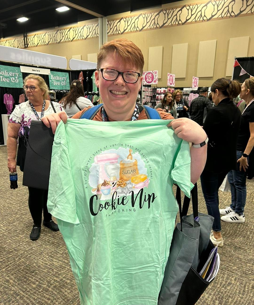 A smiling person holds up a light green t-shirt featuring a cat illustration and the text 