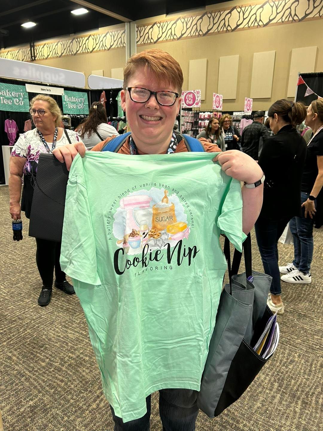 A smiling person holds up a light green t-shirt featuring a cat and the words 
