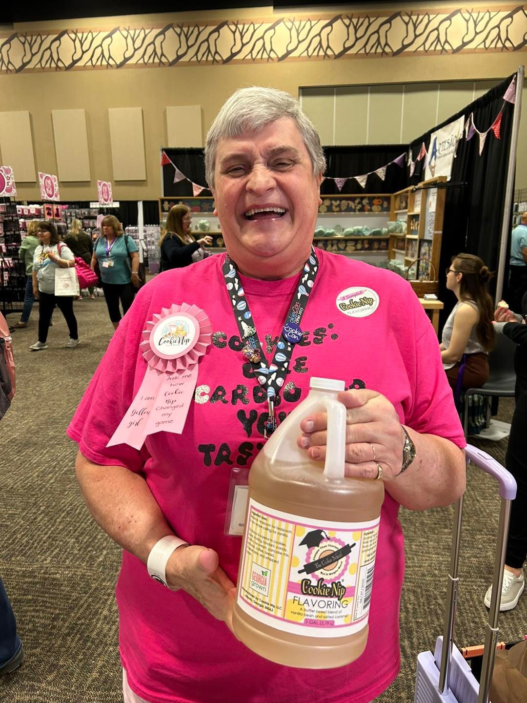 A smiling person in a pink shirt holds a large jug of Cookie Nip at a trade show or conference.