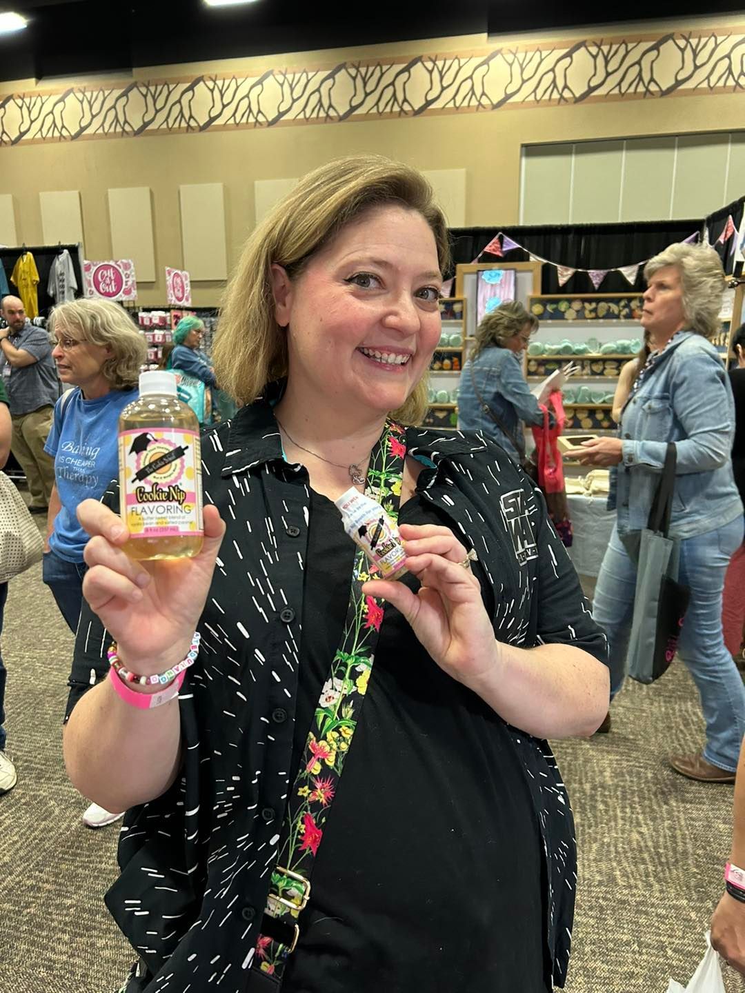 A smiling person holds a bottle of Cookie Nip in a craft market hall, wearing a patterned shirt and a lanyard.