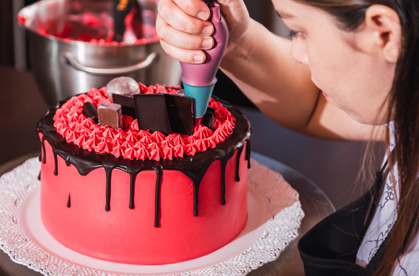 A person pipes red icing onto a bright red cake topped with chocolate decorations and a dark chocolate ganache drip.