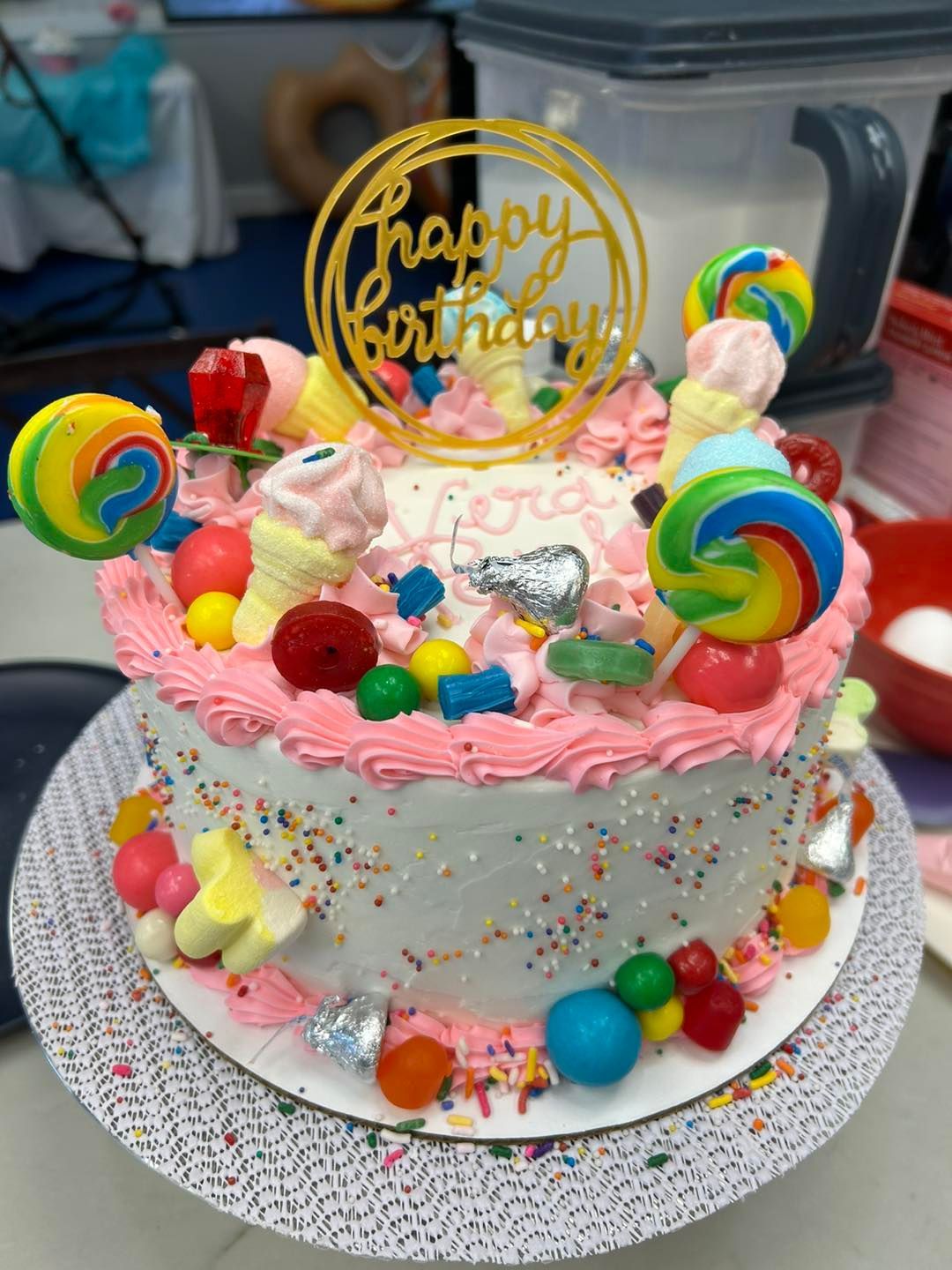A colorful, candy-topped birthday cake with white frosting, a pink border, swirls of sprinkles, and a gold cake topper.