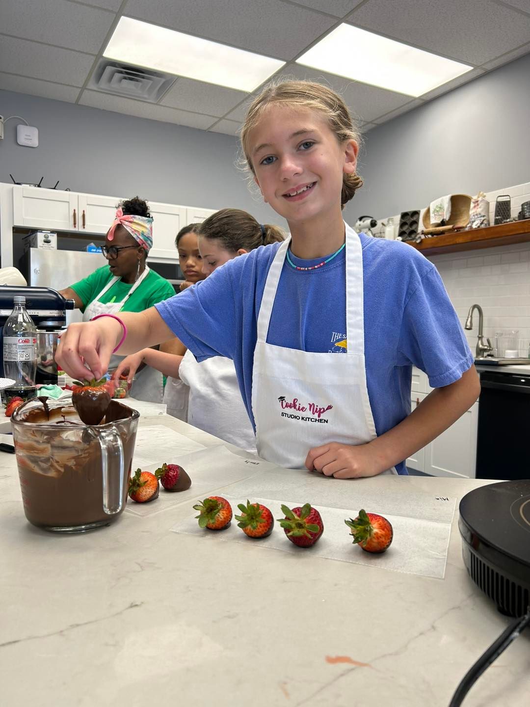A person wearing a white apron dips a strawberry into a bowl of melted chocolate in a kitchen, with more berries nearby.