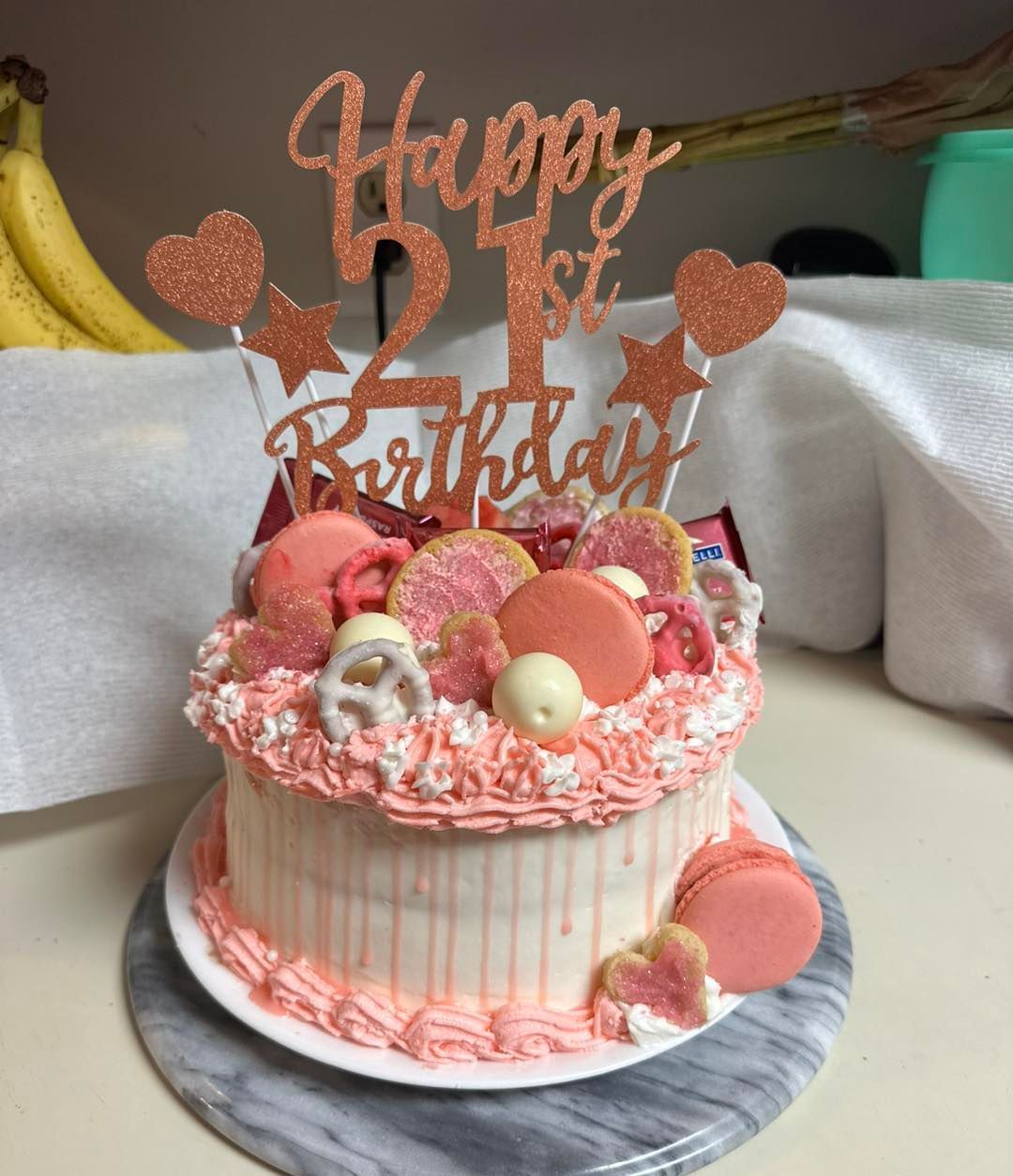 A pink, frosted 21st birthday cake topped with macarons, pretzels, and a glittery