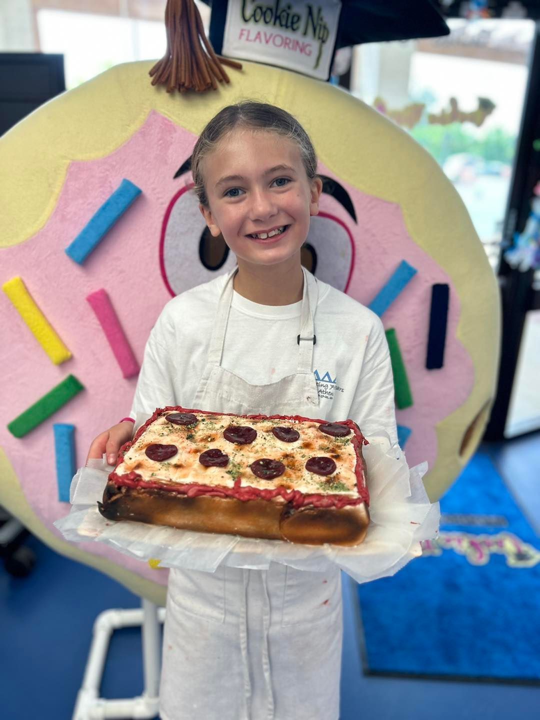 A smiling person wearing an apron holds a rectangular pepperoni pizza in front of a giant donut-shaped mascot costume.