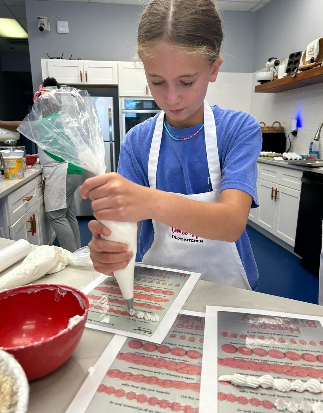 A person in a blue shirt and white apron practices piping frosting onto a printed practice sheet in a kitchen.