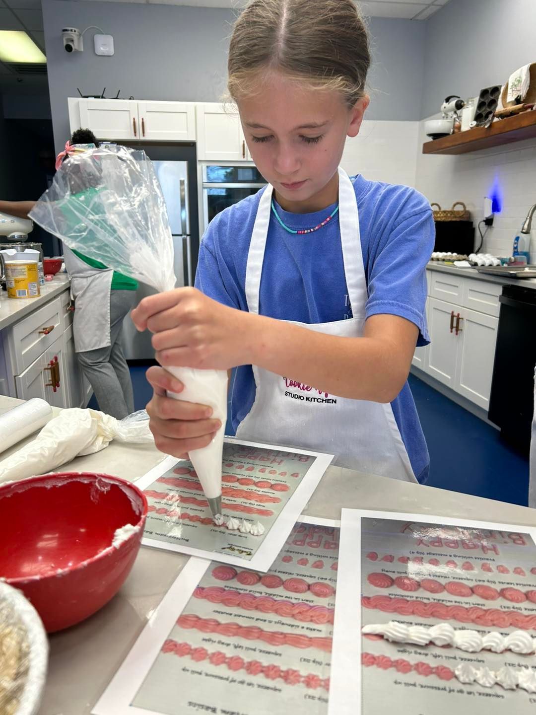 A person wearing a white apron practices piping frosting onto a template on a kitchen counter.