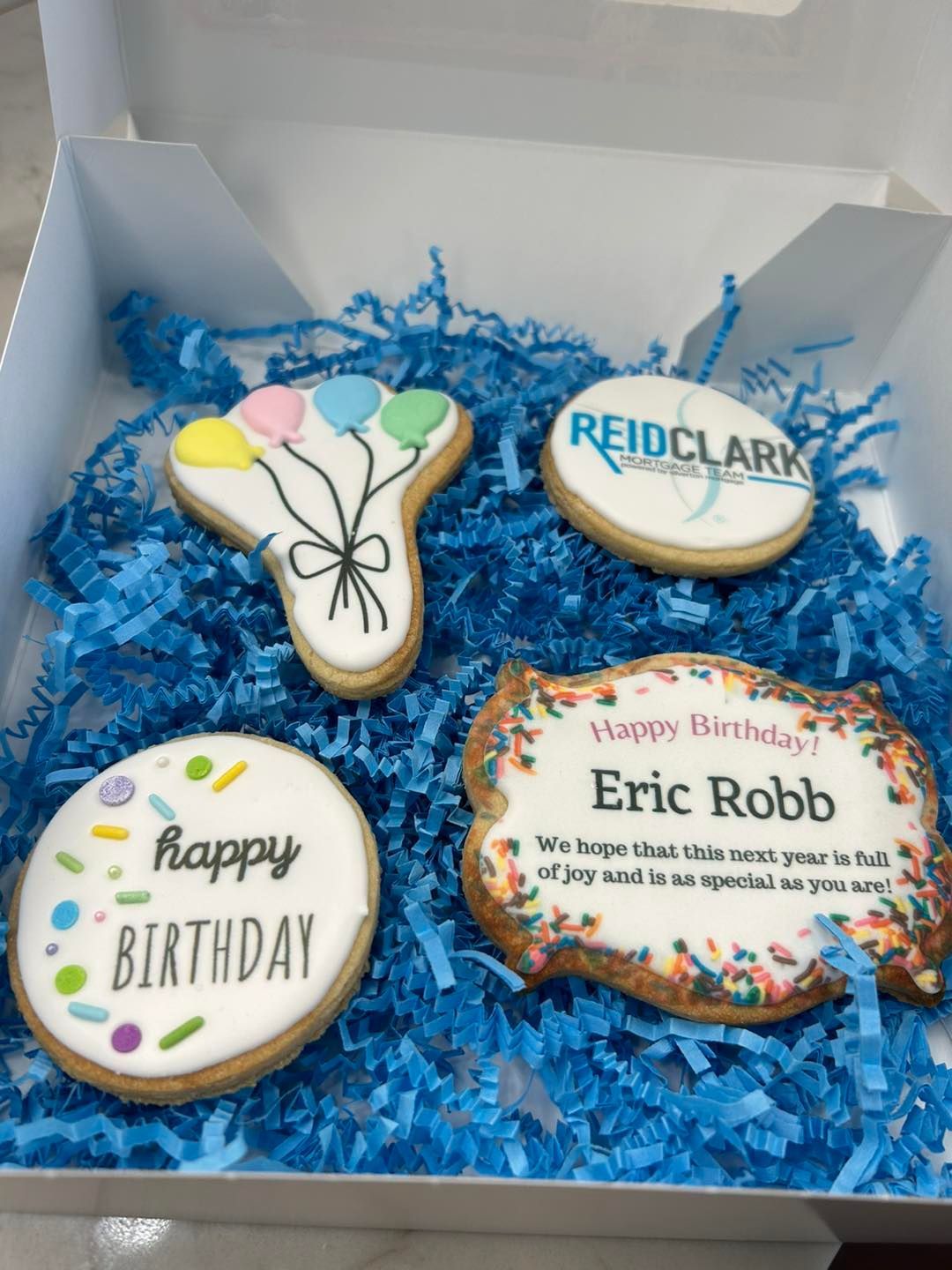 A box of four decorative birthday cookies on blue shredded paper, including one personalized for Eric Robb.