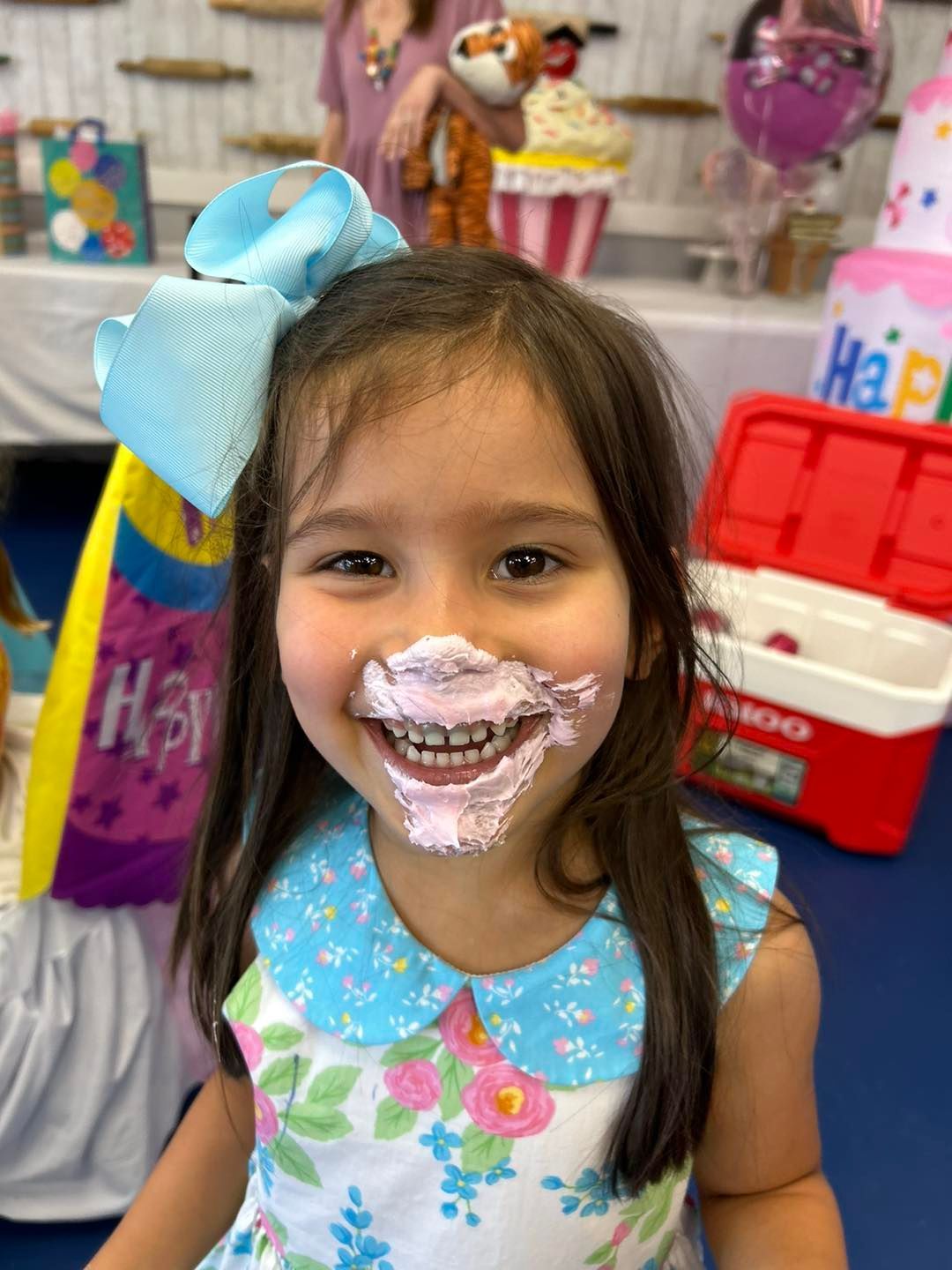 A child with frosting on their face smiles broadly, wearing a floral dress and a large blue hair bow at a birthday party.