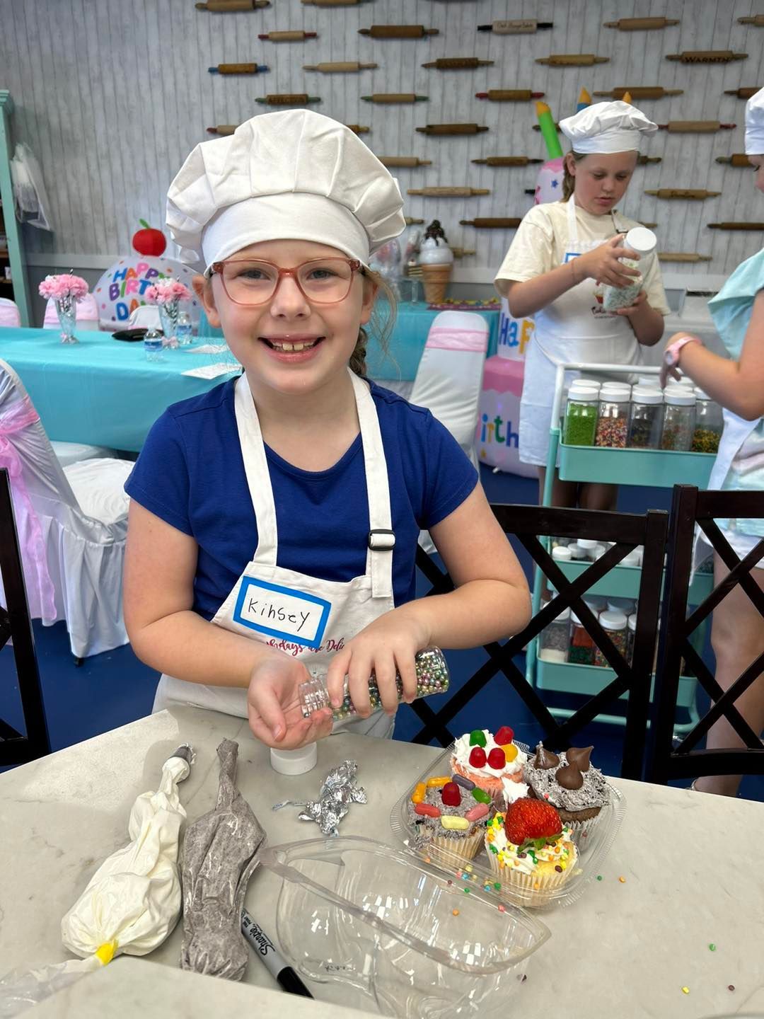 A smiling baker wears a chef’s hat and apron, decorating cupcakes with sprinkles at a craft table in a workshop setting.