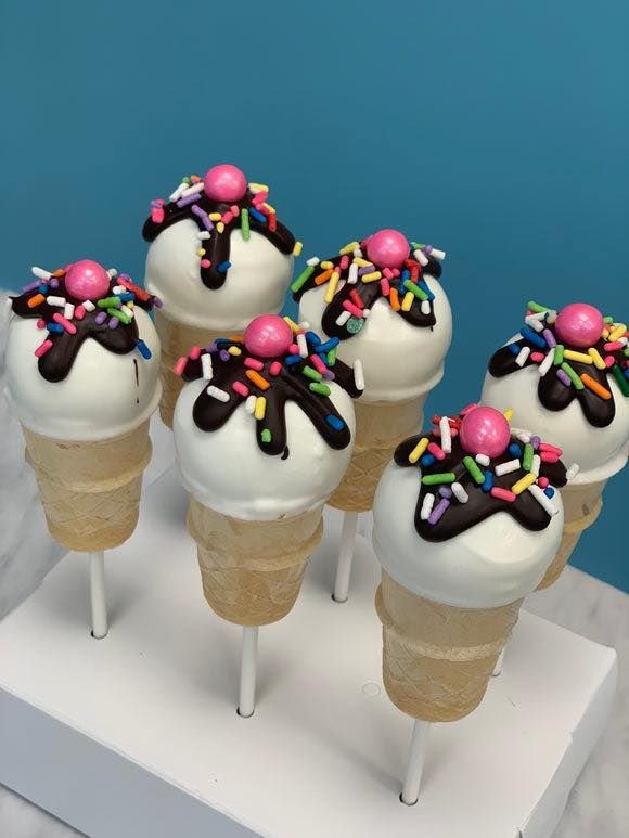 Six ice cream cone-shaped cake pops with white coating, chocolate drizzle, rainbow sprinkles, and a pink candy on top.