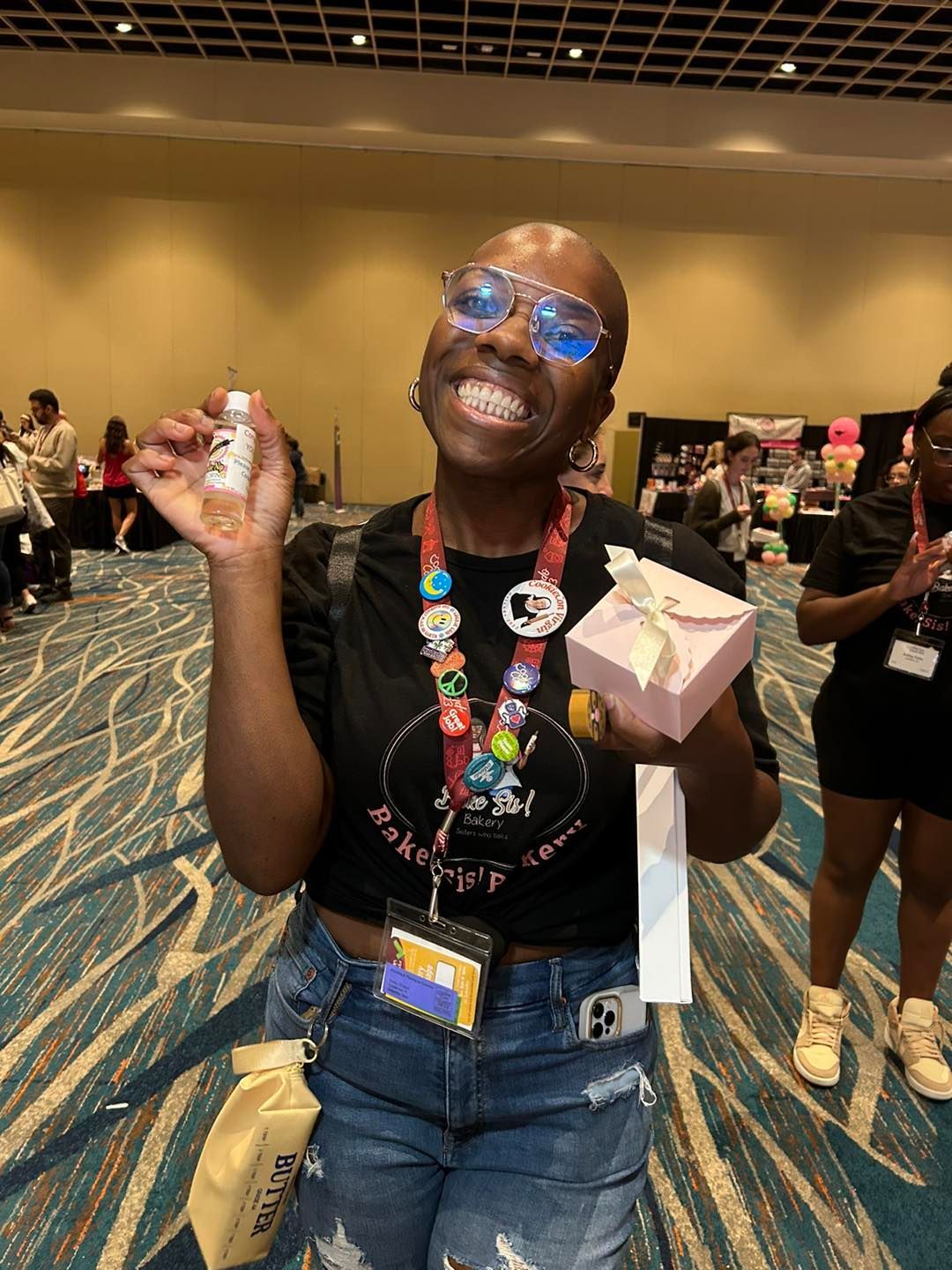 Smiling person with glasses and a lanyard holding a small bottle of Cookie Nip and a gift box in a convention hall.