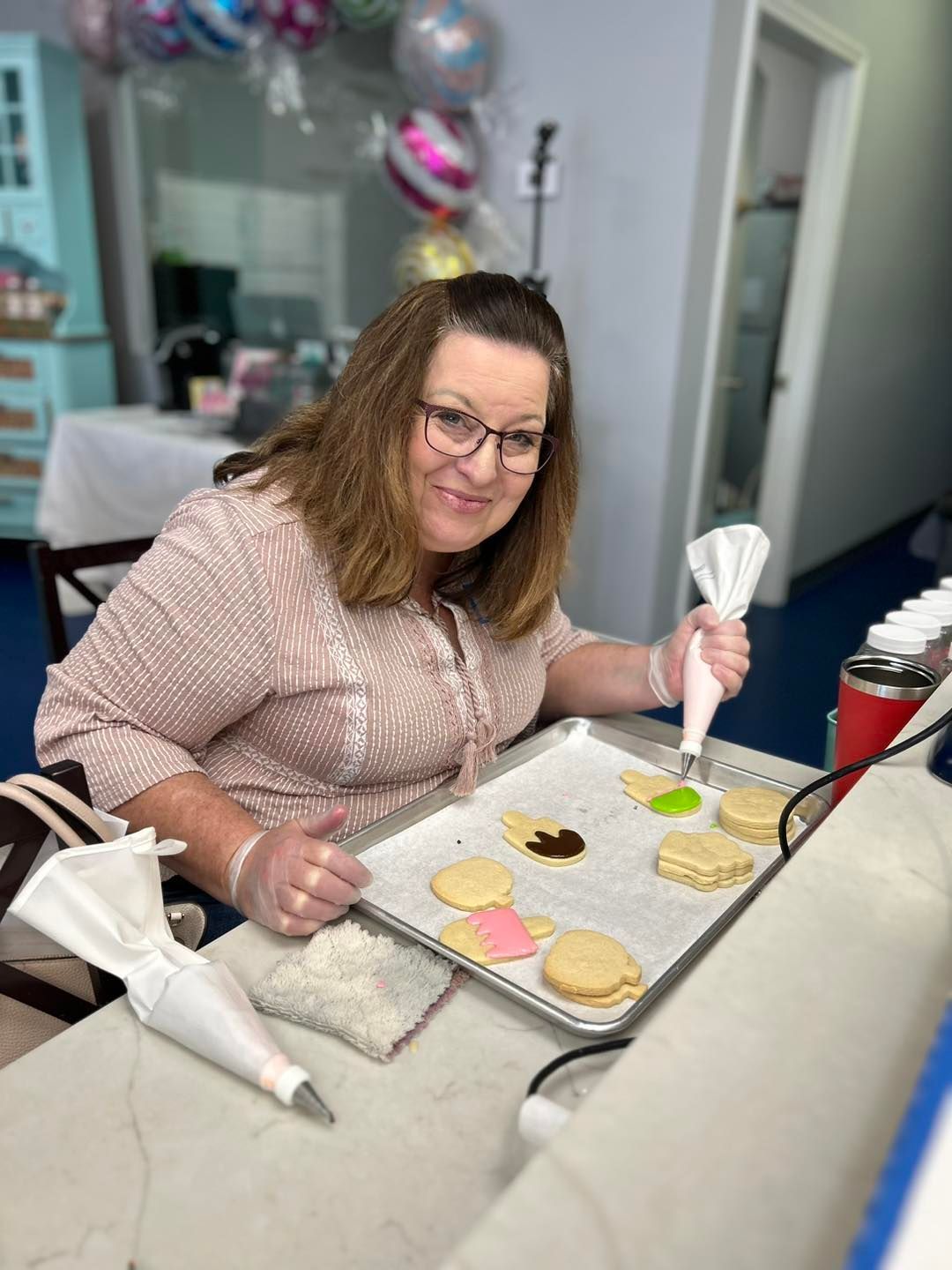 A person wearing glasses and a patterned top decorating cookies with piping bags in a brightly lit room.