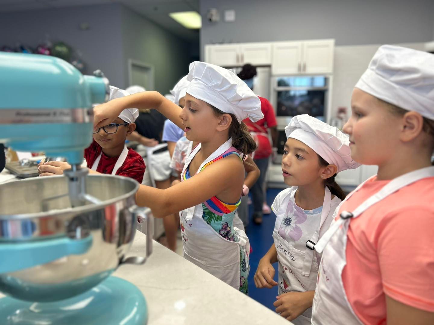 Children in chef hats and aprons, using a blue mixer in a kitchen.