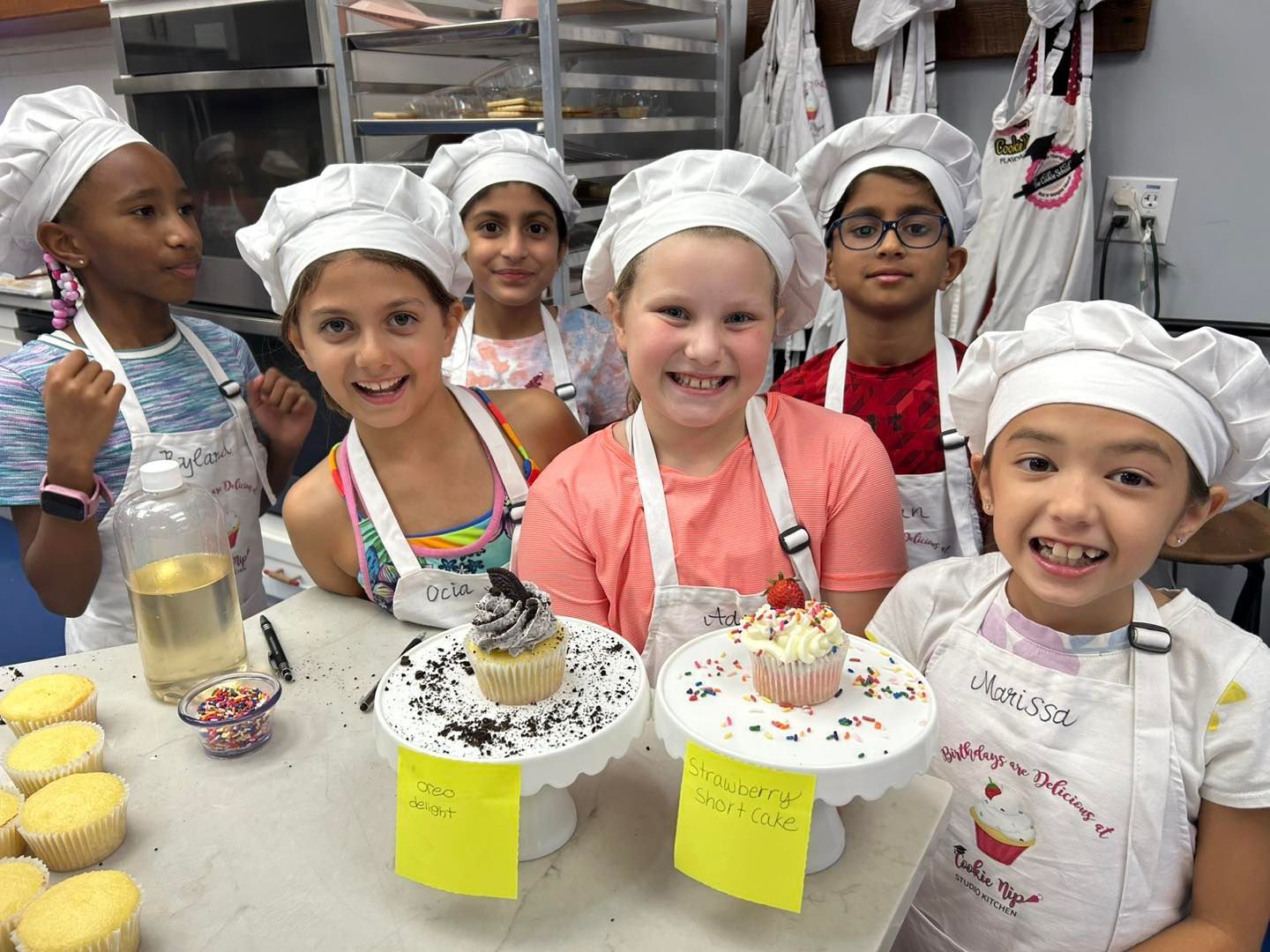Five children wearing chef hats and aprons stand smiling behind a table with cupcakes in a kitchen setting.