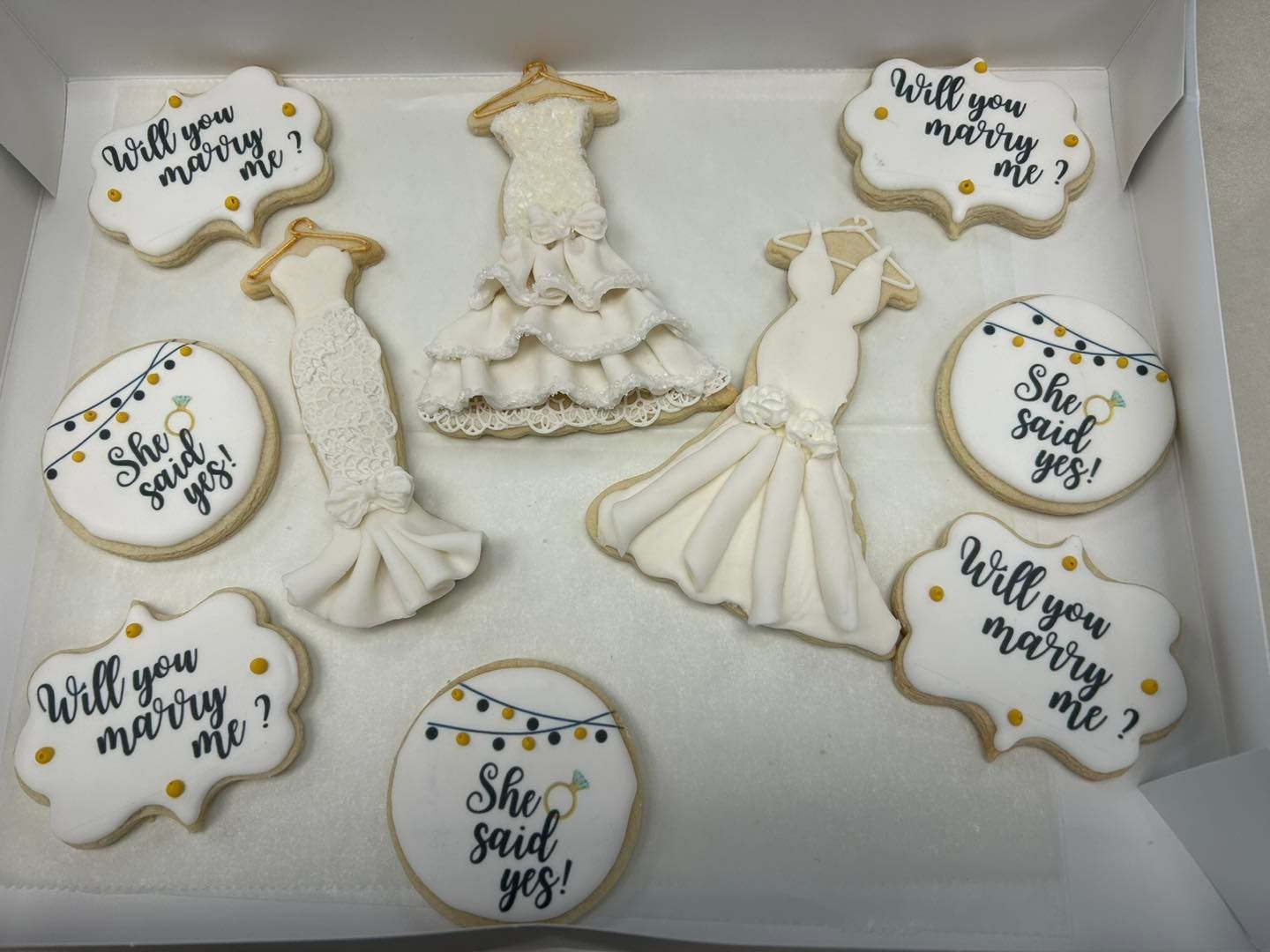 A box of wedding-themed sugar cookies featuring dress shapes and circular cookies inscribed with