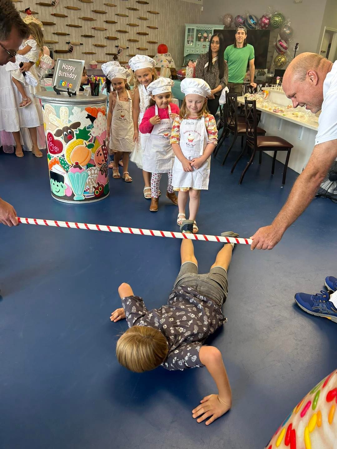 Children in aprons and hats watch a boy crawl under a held, striped pole during an indoor activity.