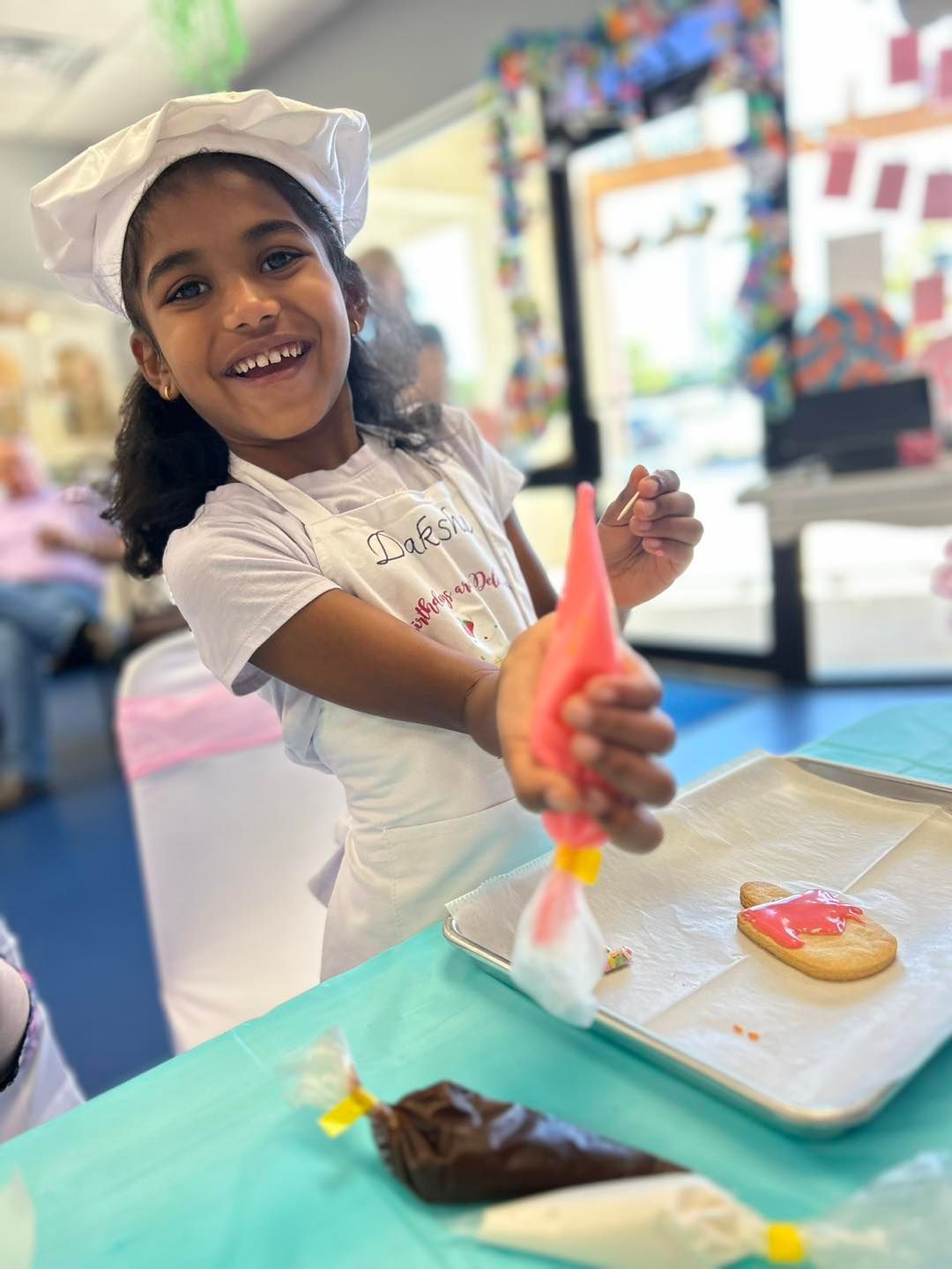 A smiling child wearing a chef hat and apron pipes pink icing onto a cookie on a tray.