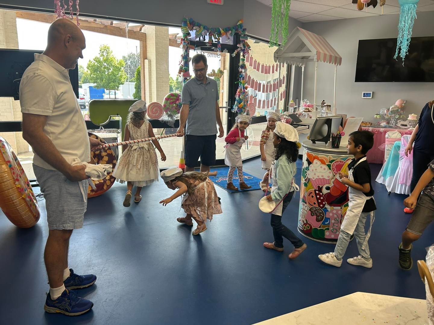 Children in aprons and hats participate in a party activity at an indoor venue while two adults supervise.