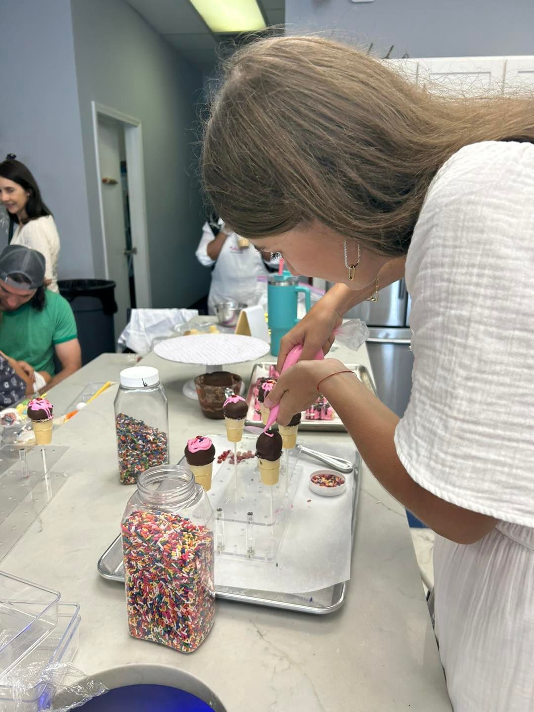 A person uses a pink piping tool to decorate chocolate-dipped ice cream cones on a tray at a table with sprinkles.