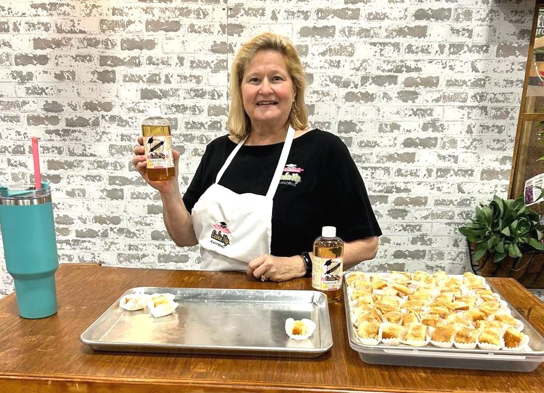 A smiling woman in an apron stands behind a wooden table with trays of small pastries and bottles of liquid.