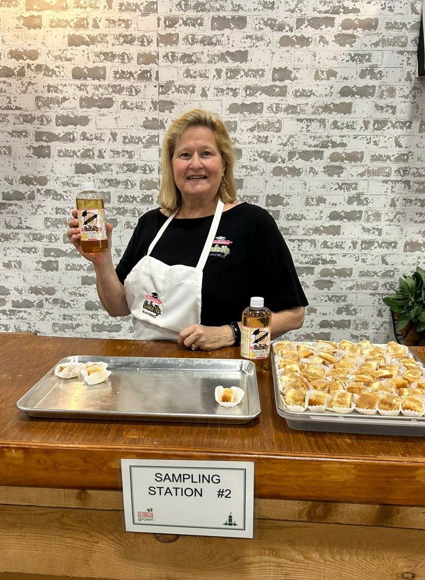 A smiling person in an apron stands at a sampling station with trays of food and bottles of Cookie Nip against a brick wall.