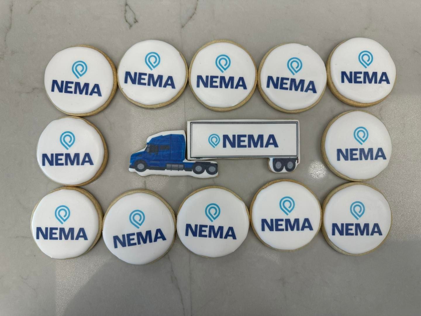 Twelve circular cookies with the NEMA logo arranged around a central truck-shaped cookie on a light marble surface.