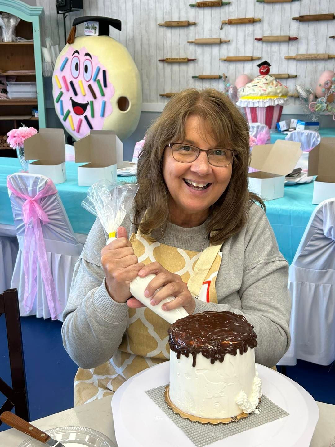 A person smiles while piping icing onto a white-frosted cake with a chocolate drip in a room decorated with baking items.