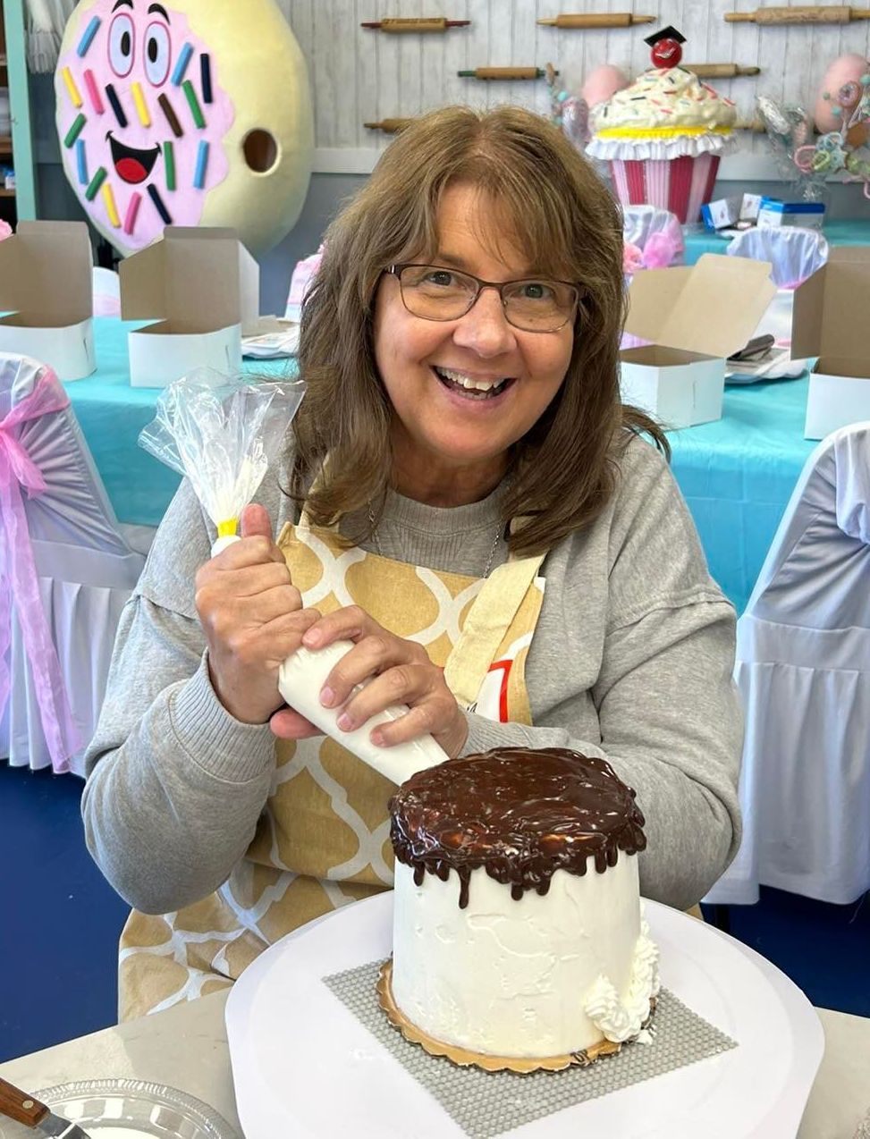 A person with brown hair and glasses smiles while piping white frosting onto a small cake topped with chocolate ganache.