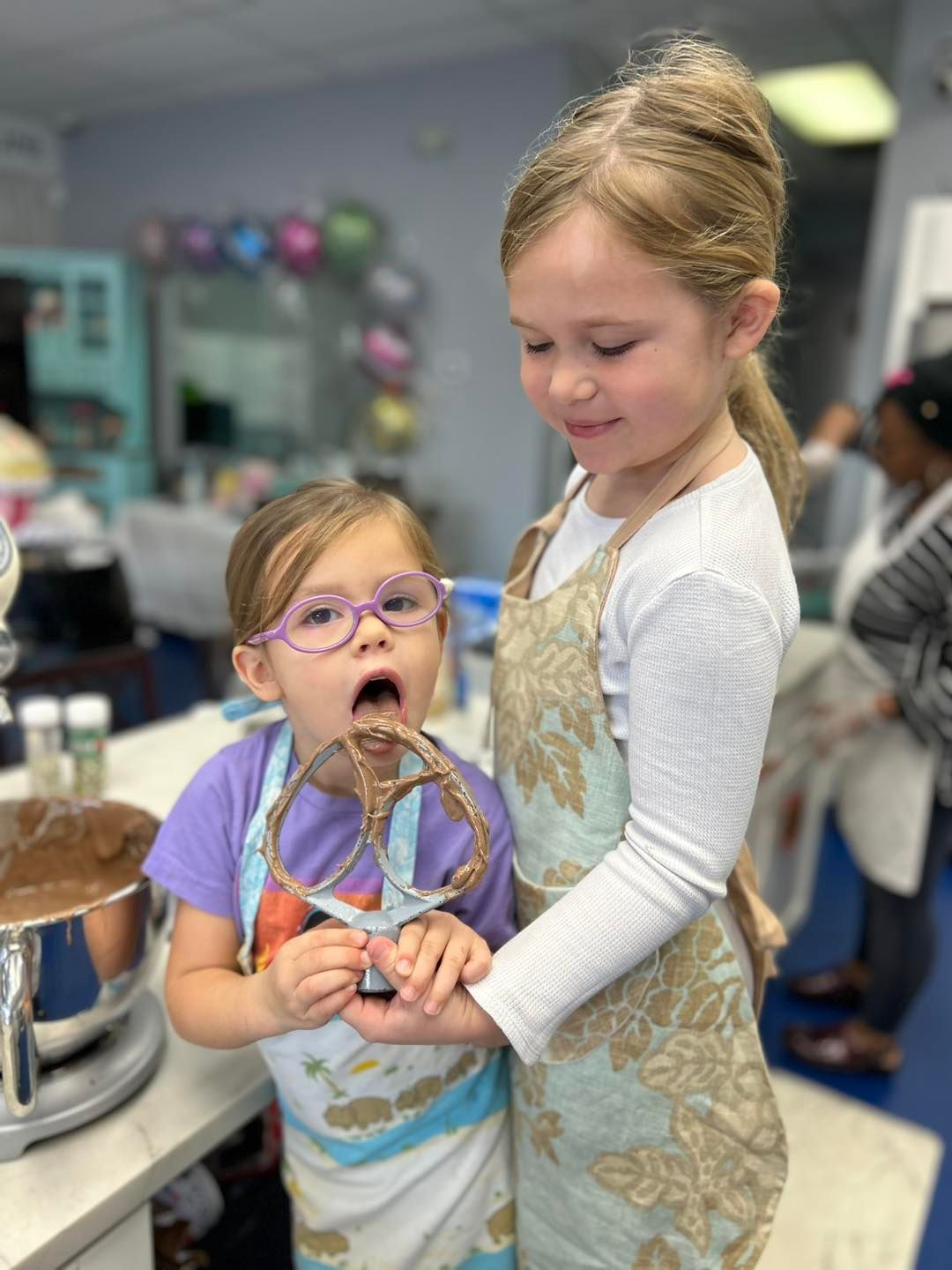 Two children in aprons stand in a kitchen; one licks chocolate off a metal mixer attachment, the other holds it steady.
