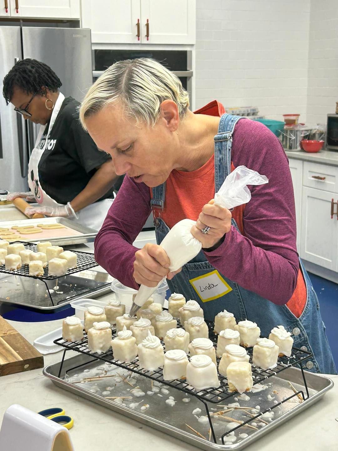 A person wearing an apron pipes icing onto small, square cakes arranged on a cooling rack in a bright kitchen.