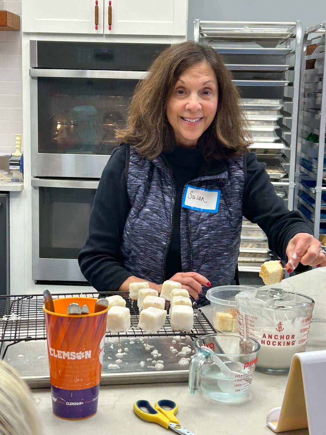A person in the Cookie Nip Studio Kitchen glazes square treats on a wire rack, with baking equipment