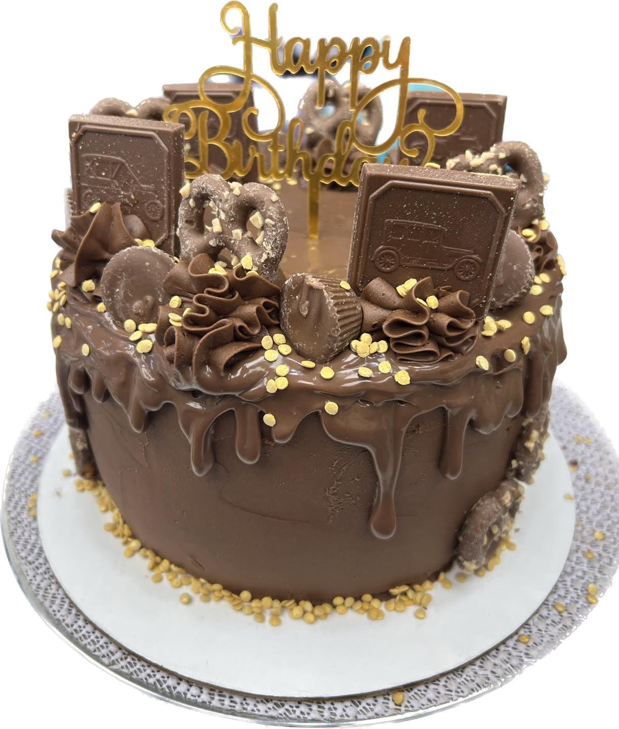 A round, brown chocolate birthday cake topped with chocolate squares, pretzels, peanut butter cups, and gold sprinkles.