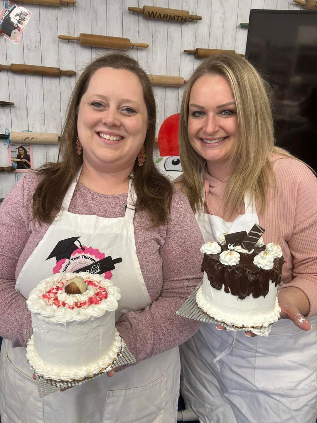 Two people wearing aprons hold small decorated cakes in a kitchen with wooden rolling pins on the wall behind them.