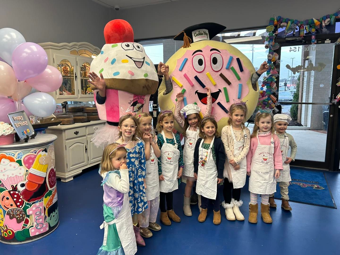 Children in white aprons stand with large cupcake and donut mascots in a bakery shop decorated for a party.