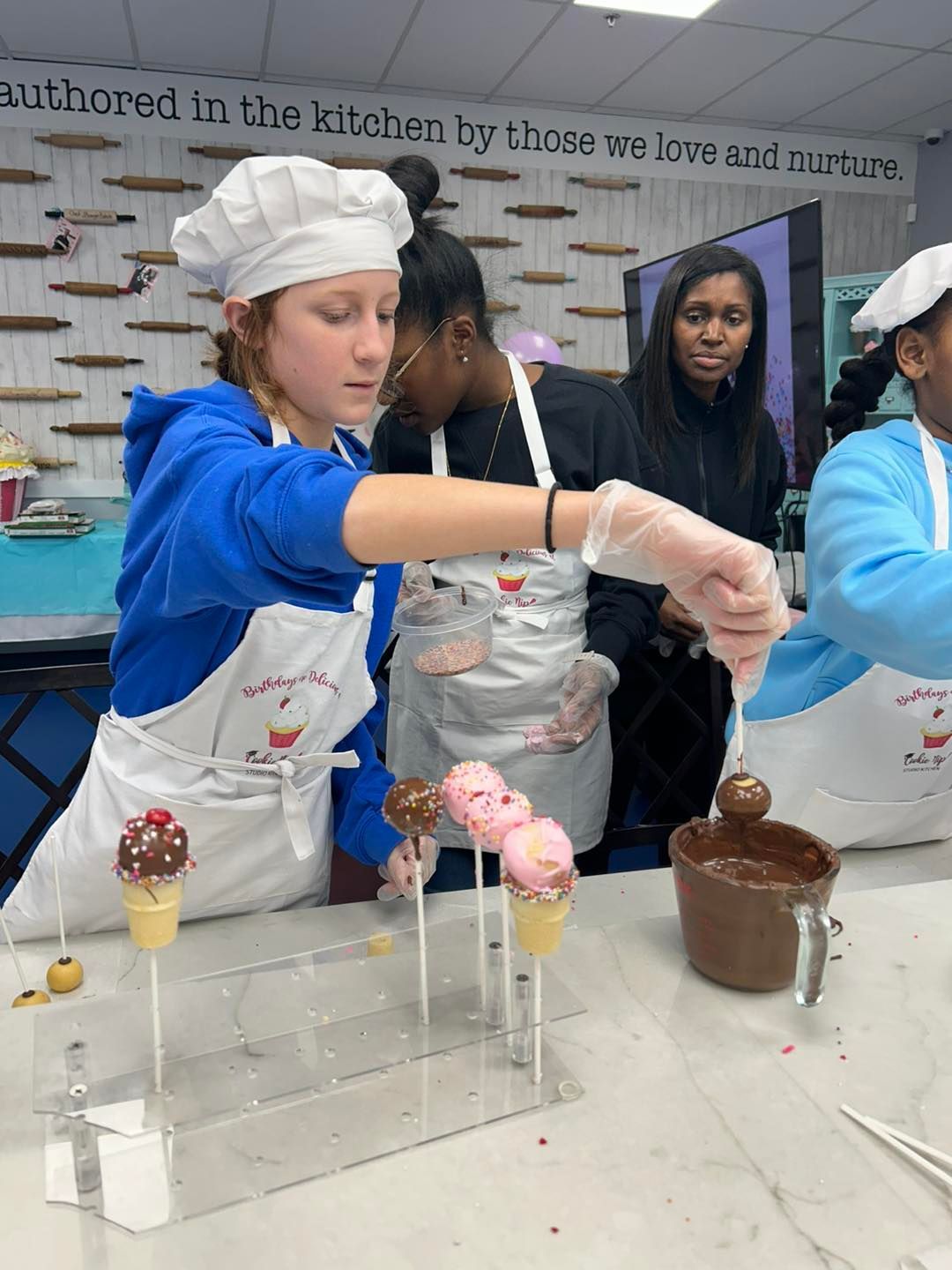 People in aprons decorate cake pops by dipping them into a container of melted chocolate in the Cookie Nip Studio Kitchen.