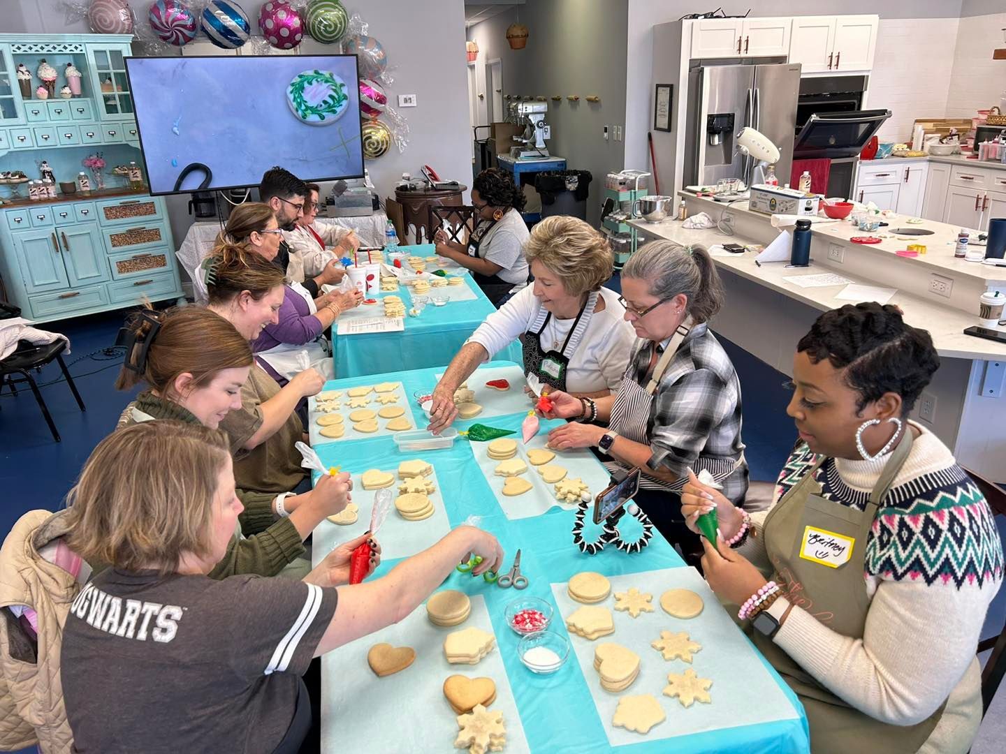 A group of people seated at tables in a bright kitchen, decorating sugar cookies with icing and sprinkles.