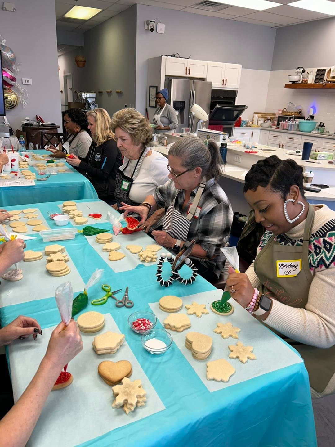 A group of people decorates cookies with icing at a table covered with a teal tablecloth during a baking class.