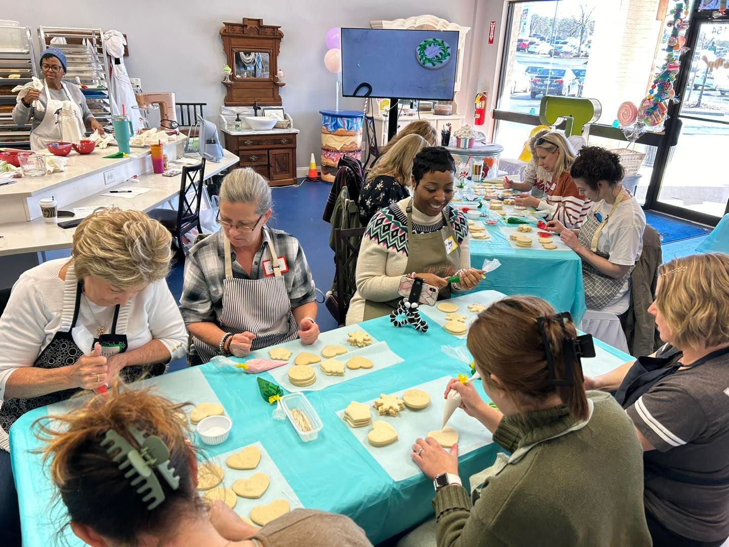 A group of people decorates cookies together around a table covered in a bright blue tablecloth in a well-lit workshop.
