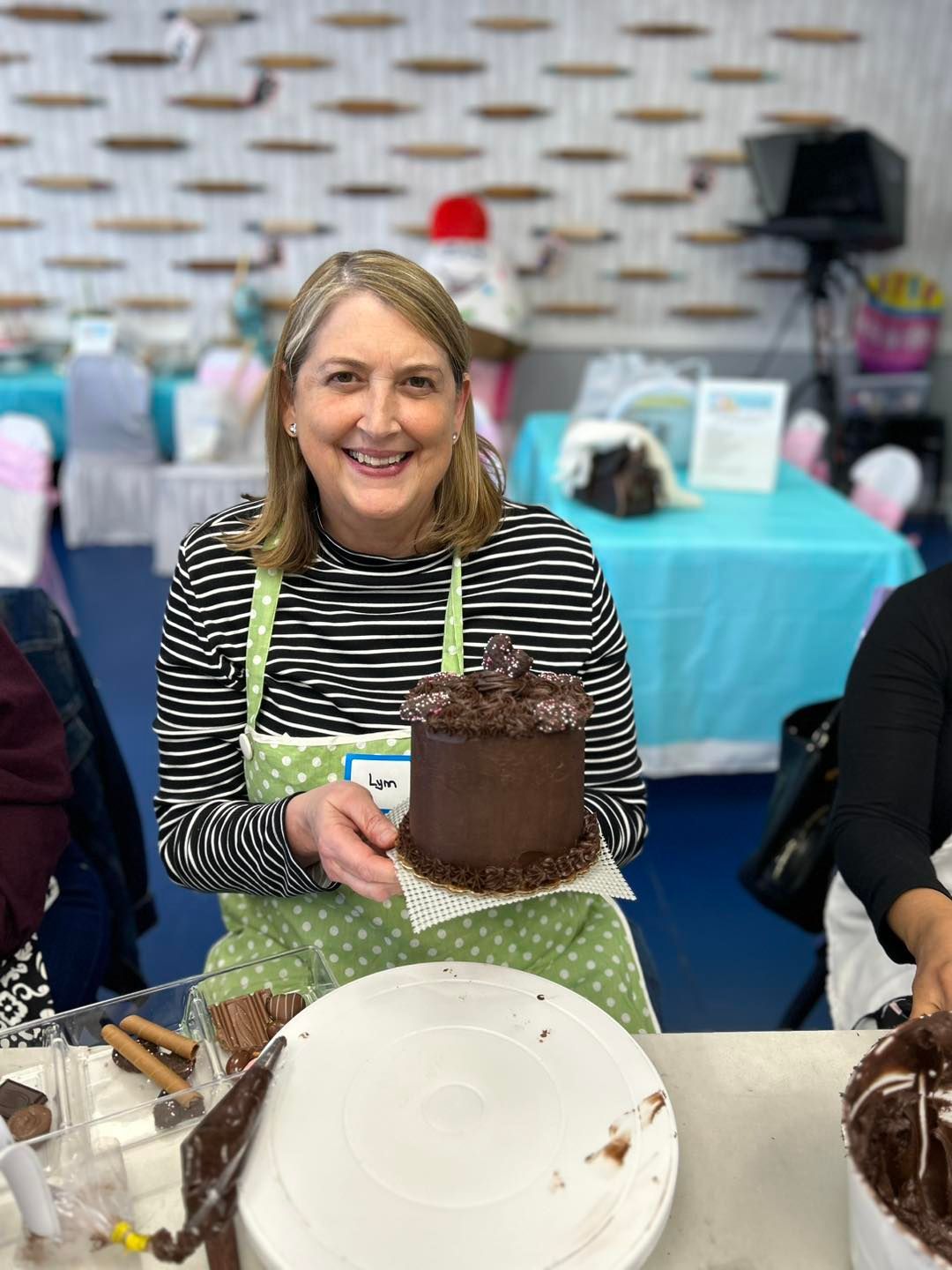A smiling person in a green polka-dot apron holds a small, round, dark chocolate cake in a bright, decorated room.