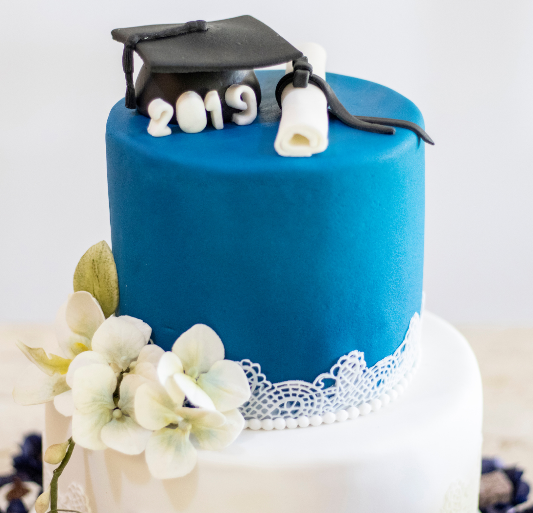 A two-tier graduation cake with a blue top, white bottom, white lace trim, fondant flowers, a cap, and a diploma.