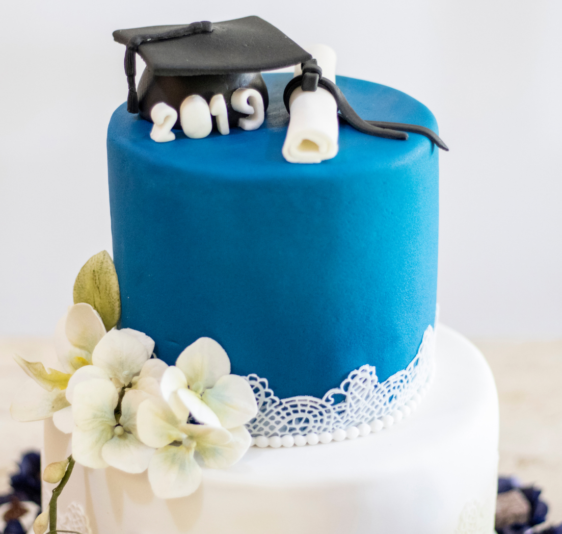 Two-tiered graduation cake with blue fondant, a black cap topper labeled 2019, a diploma, and white lace and floral accents.