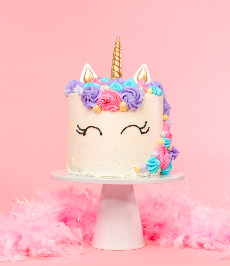Unicorn-themed cake with floral frosting, gold horn, and closed eyes on a pedestal, surrounded by pink feathers.