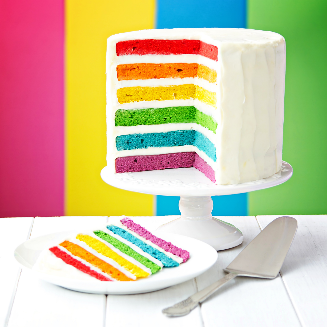 A rainbow-layered cake with white frosting sits on a stand, with a slice on a plate and a cake server on a white surface.