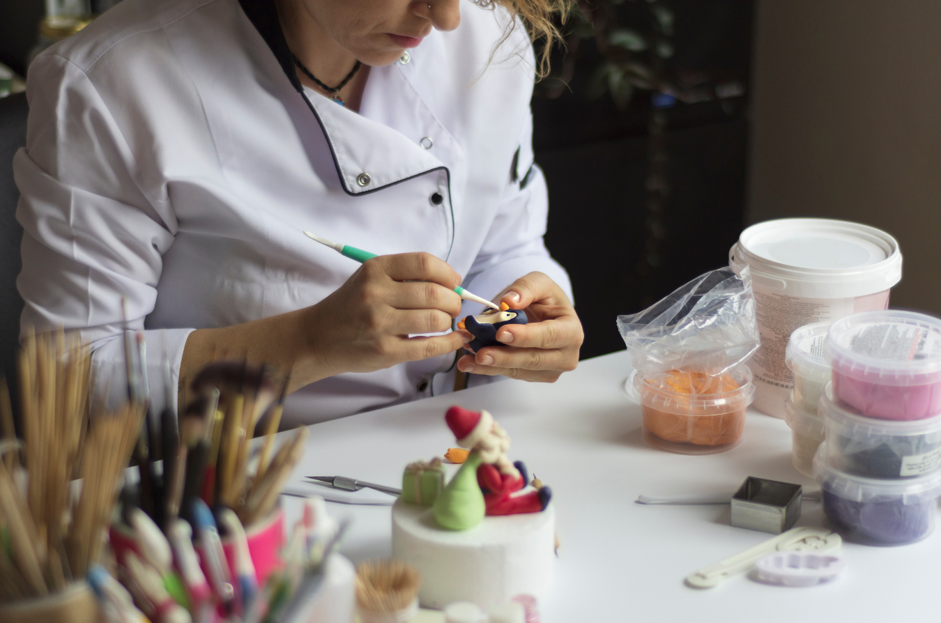 A baker in a white uniform works on a small fondant Santa figurine at a table with tools and containers of supplies.