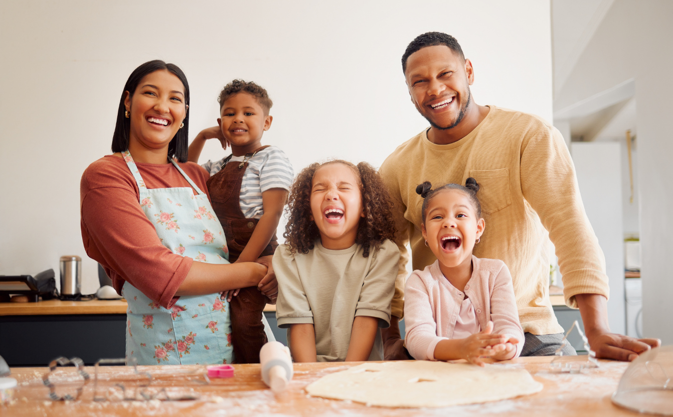 A family laughing while baking cookies together in a kitchen, with dough and cookie cutters on the counter.