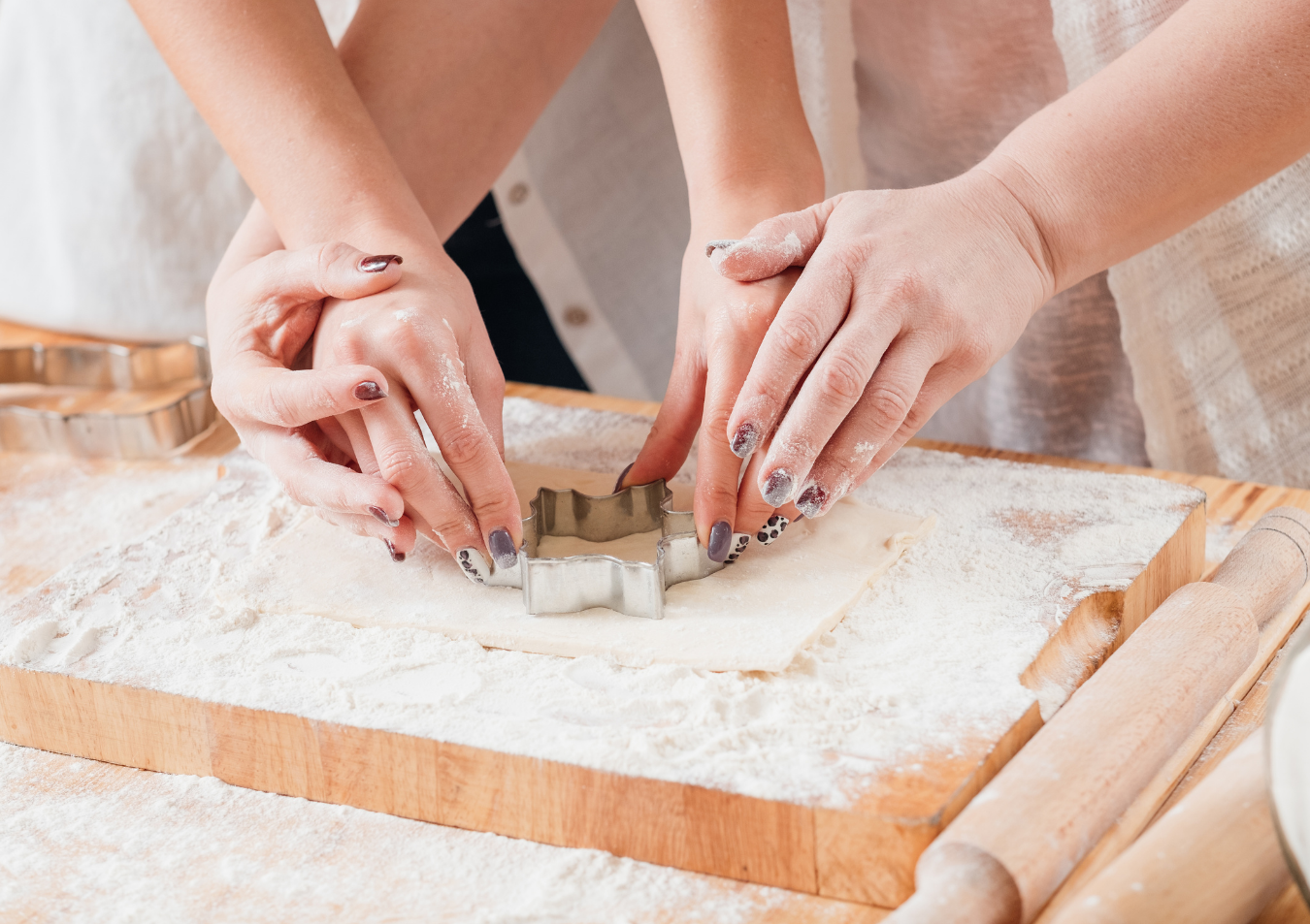 Two pairs of hands pressing a metal cookie cutter into rolled-out dough on a floured wooden surface.